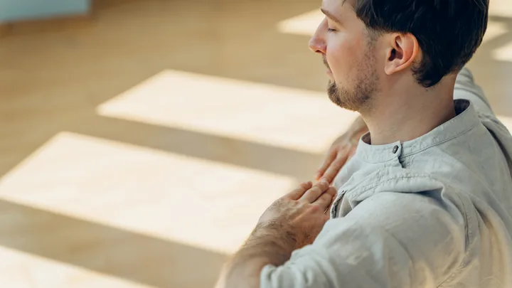 A man breathing slowly in a sunlit room, one hand resting on his chest.