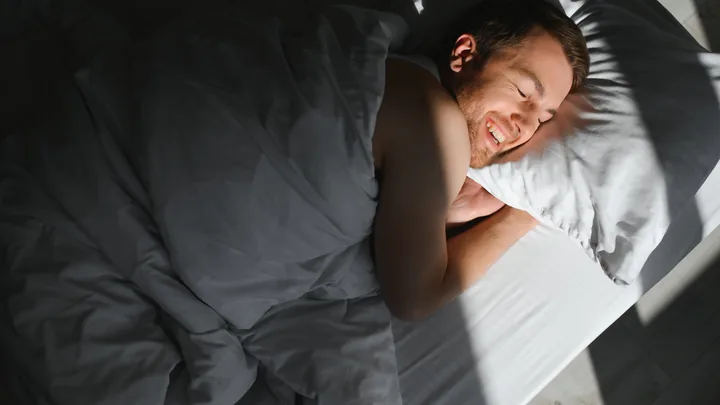 A man waking slowly in a bed lit by morning sun through the blinds.