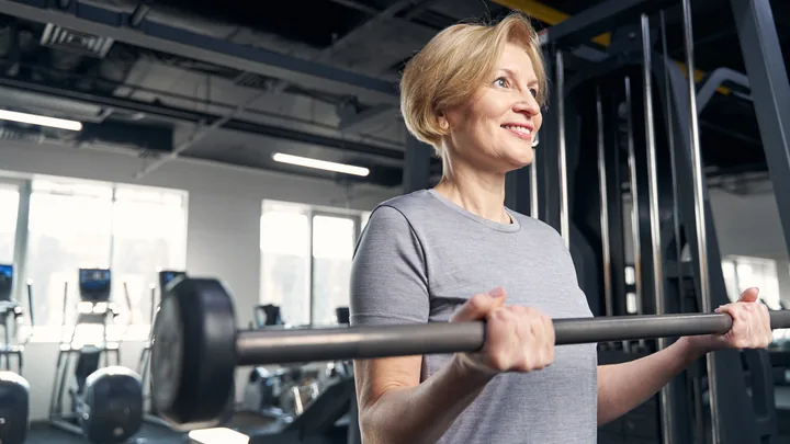 A smiling woman holding a loaded barbell across her shoulders in a bright gym.