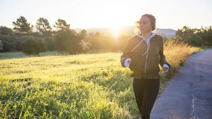 A woman running along a rural road through tall grass at sunrise.