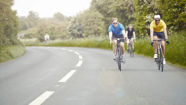 Three cyclists riding at an easy pace along a sunny country road lined with trees.
